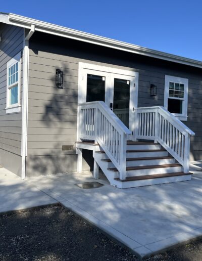 A gray house with white trim, featuring a small porch and stairs leading to a glass door. The yard is partially paved.