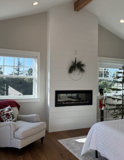 A cozy bedroom with a fireplace, armchair, and bed. Large windows let in natural light. Decor includes a small tree and wreath.