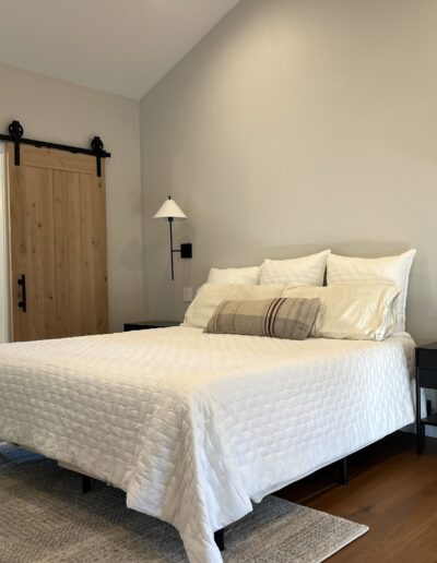Modern bedroom with a white bedspread, wooden sliding barn door, black nightstands, and wall-mounted lamps. A large window is visible in the adjacent room.