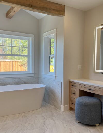 A bathroom with a freestanding bathtub, a wooden vanity with a mirror, a cushioned stool, and a window with a garden view.