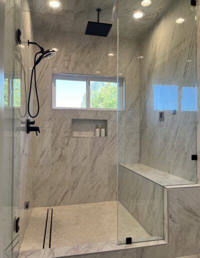 Modern glass-enclosed shower with marble walls, black fixtures, and a built-in bench. There are two bottles on a small shelf and a window outside.